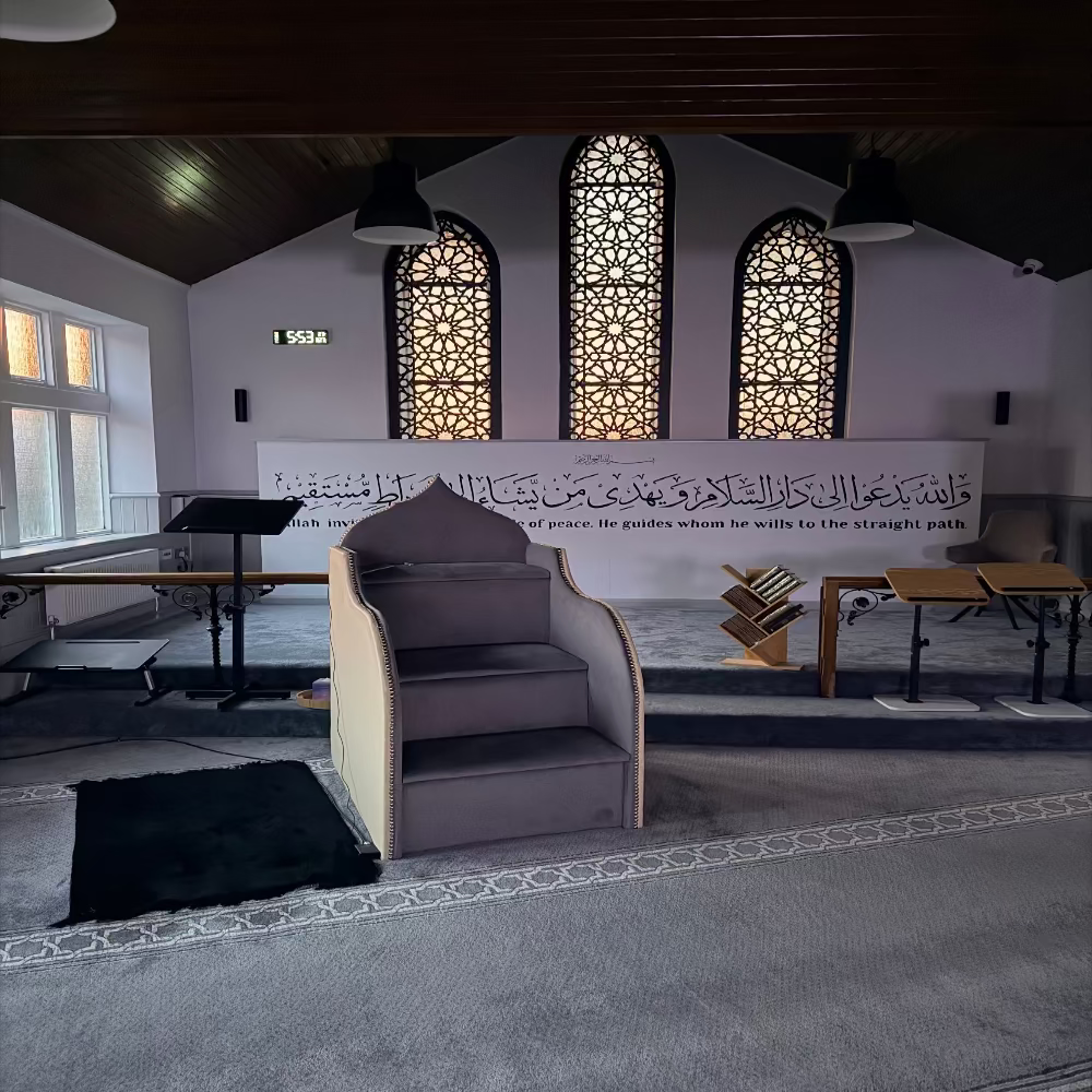 Interior of the Masjid with a large wall inscription and a prayer mat on the floor.