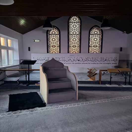 Interior of the Masjid with a large wall inscription and a prayer mat on the floor.